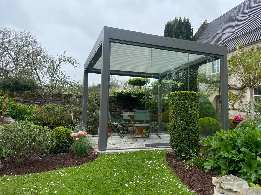 Pergola bioclimatique anthracite, lames claires, parois vitrées, jardin pierre et pelouse