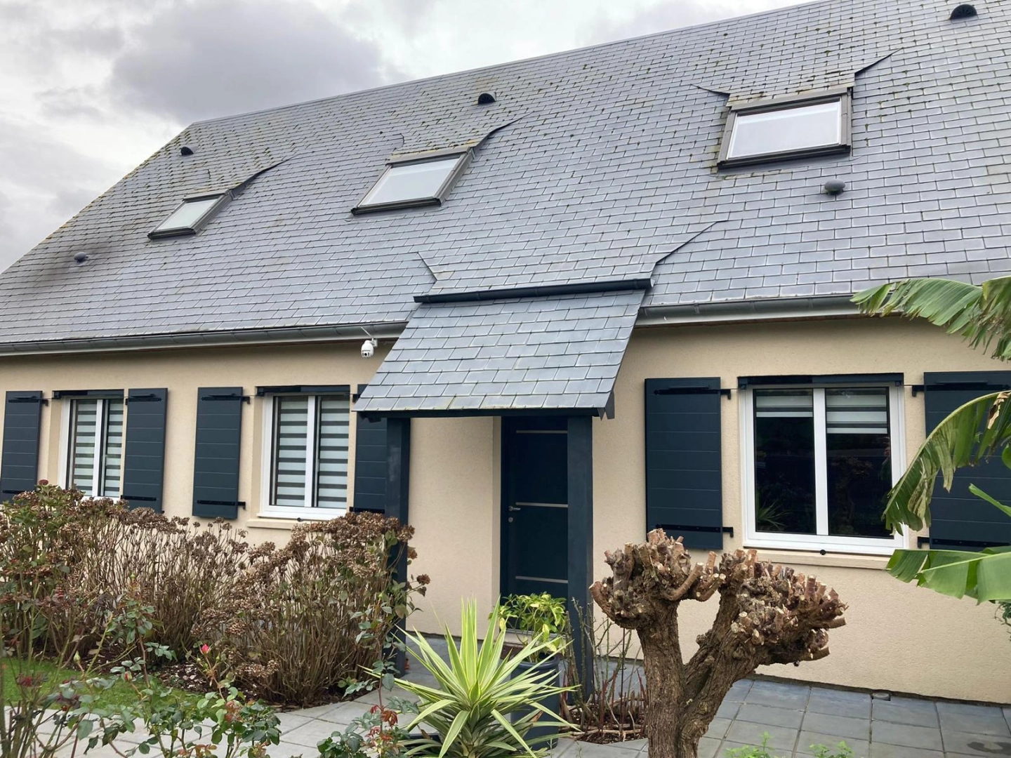 Façade crème, volets battants anthracite, porte sous auvent ardoise, velux toit