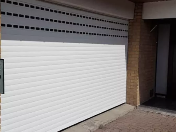Porte de garage enroulable blanche, trois rangées de hublots noirs, piliers briques beige