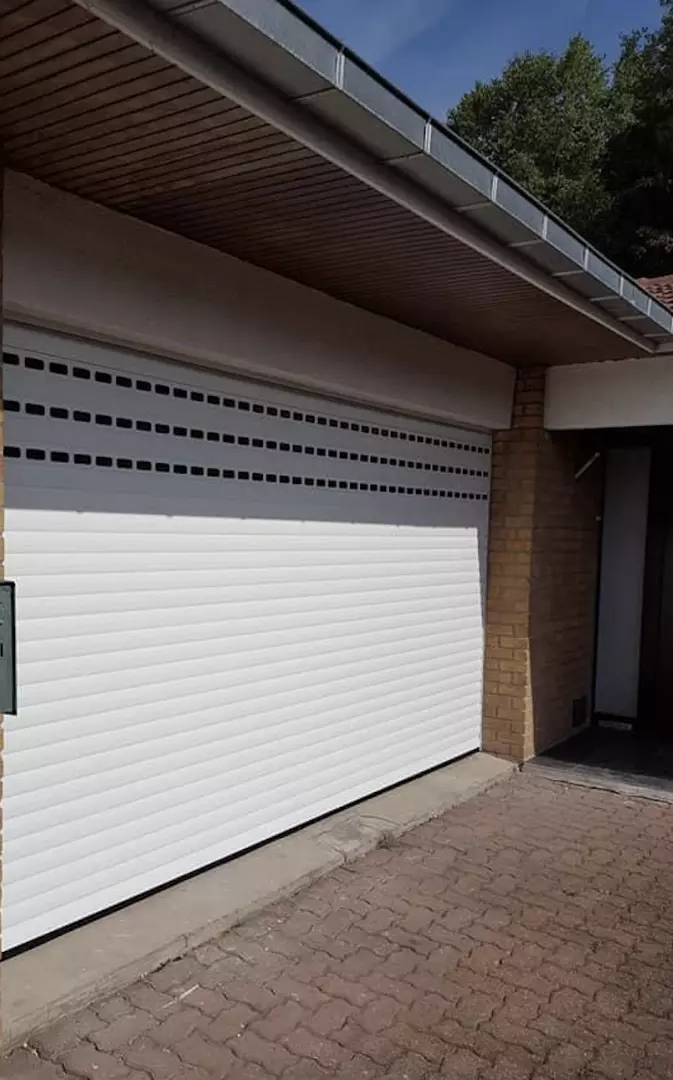 Porte de garage enroulable blanche, trois rangées de hublots noirs, piliers briques beige