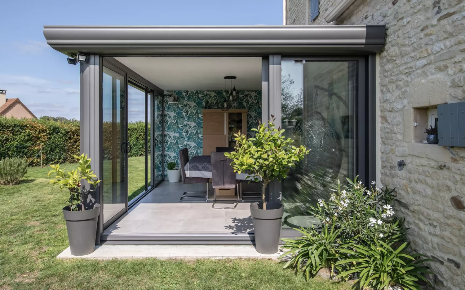 Véranda toit plat alu gris architecte, baies coulissantes, maison pierre et pelouse