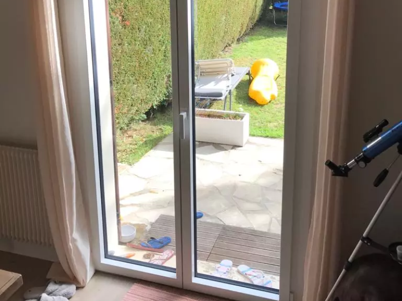 Porte-fenêtre PVC blanche deux vantaux, vue terrasse pierre, jardin et haie verte
