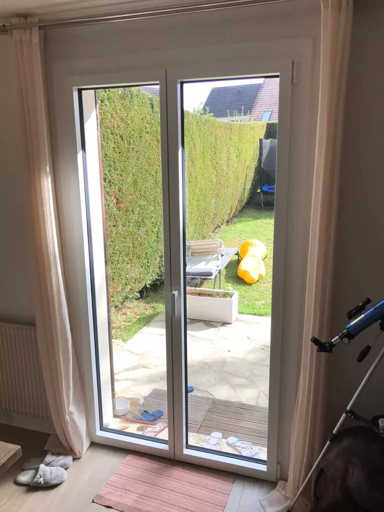 Porte-fenêtre PVC blanche deux vantaux, vue terrasse pierre, jardin et haie verte