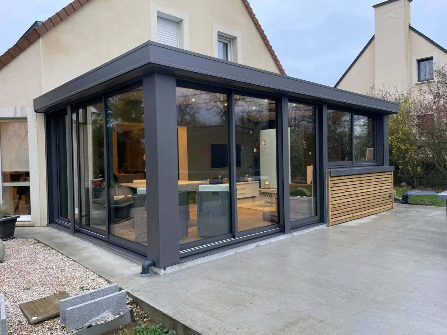 Véranda anthracite toit plat, baies coulissantes et bardage bois clair sur maison tuiles brunes