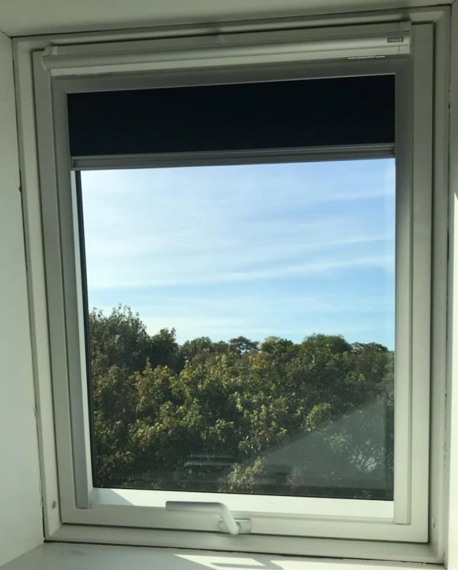 Fenêtre blanche encastrée, store occultant gris mi-baissé, ciel bleu et cimes d'arbres