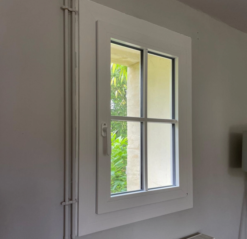 Fenêtre PVC blanche quatre carreaux, poignée gauche, vue feuillage et mur pierre