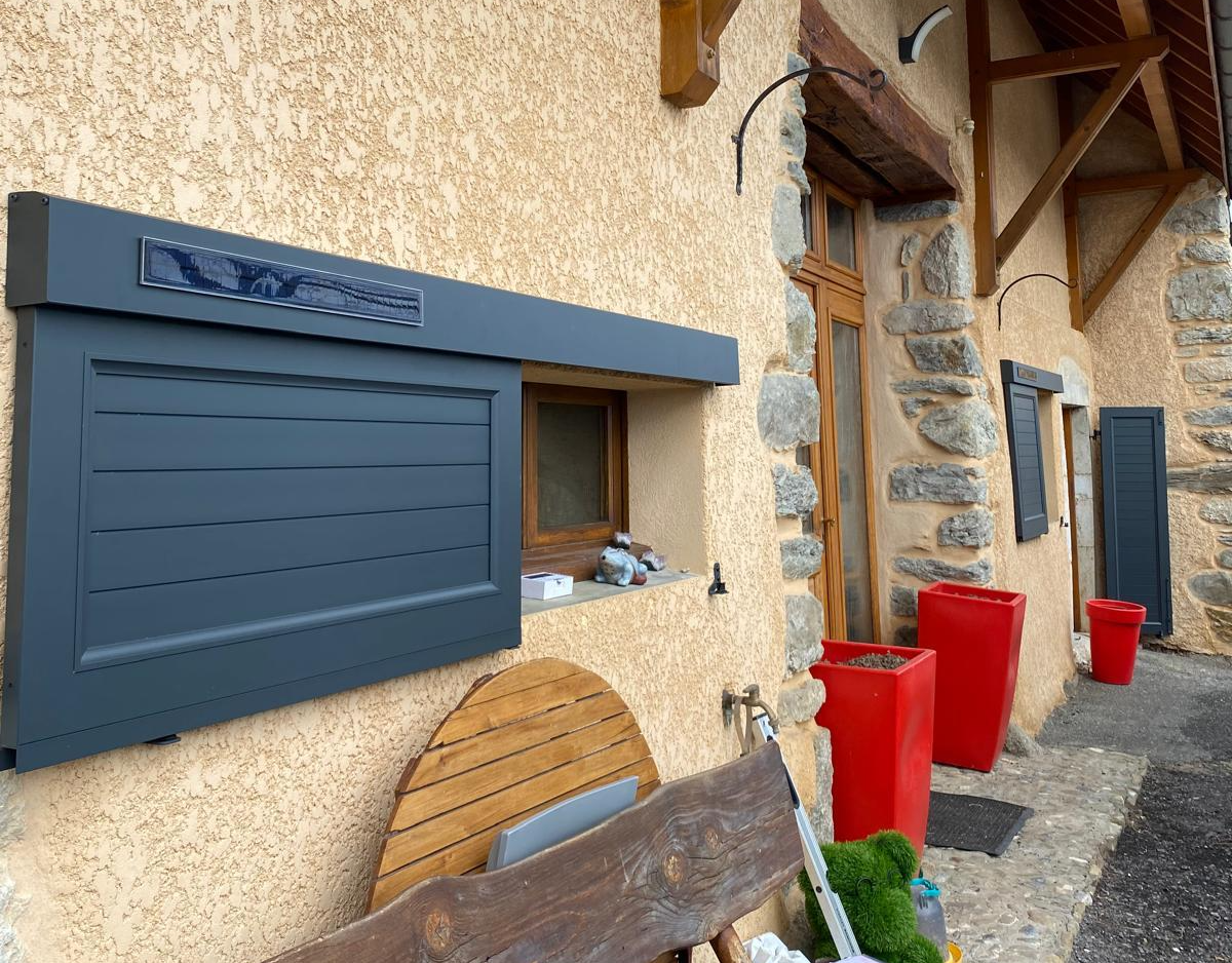 Volet coulissant anthracite solaire à lames sur mur crépi, banc en rondins et jardinières rouges