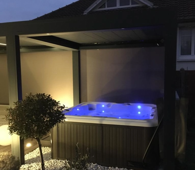 Spa carré éclairage bleu sous pergola noire, galets blancs et jardin au crépuscule