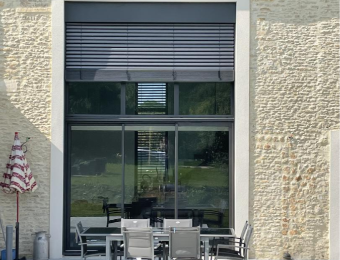 Baie coulissante anthracite sur mur pierre, BSO gris partiellement baissé, terrasse et table