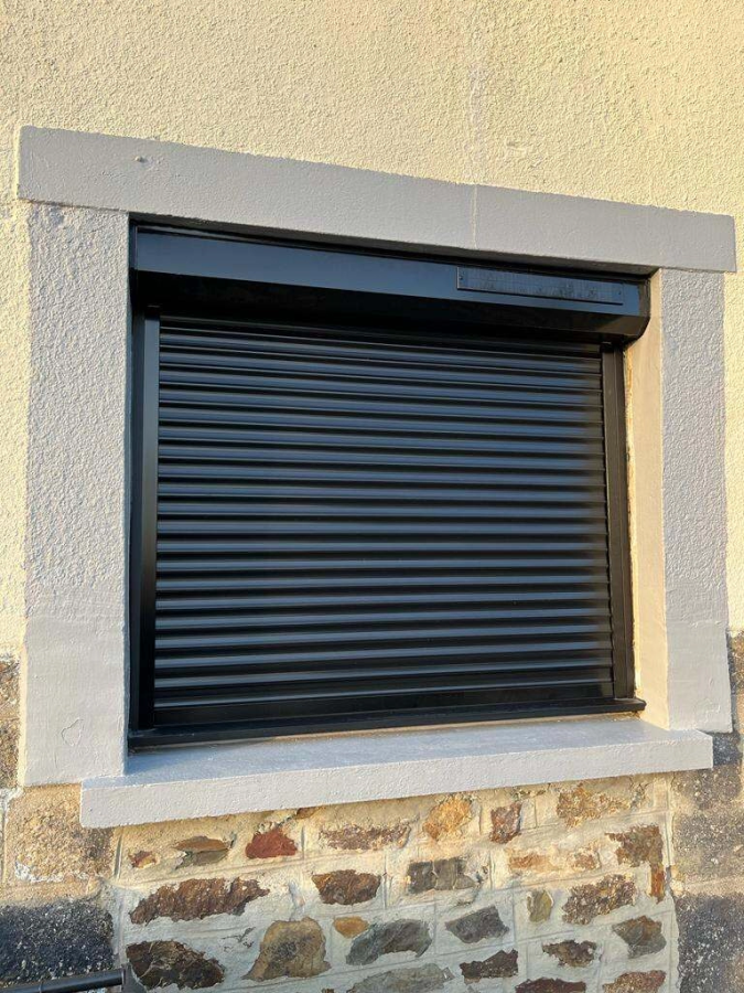 Volet roulant noir fermé, panneau solaire sur coffre, encadrement gris, mur pierre