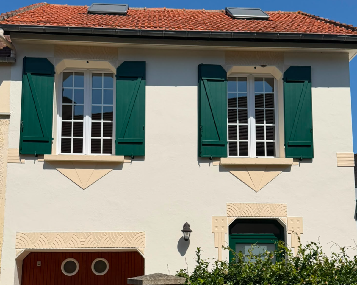 Maison crépi clair, volets battants verts ouverts aux fenêtres d’étage, porte garage bois et panneau Logikinov